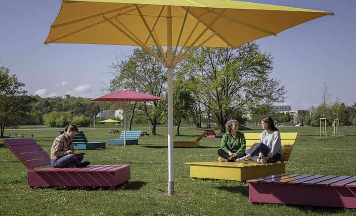 großer gelber Sonnenschirm und pinker Schirm über pinken und gelben Außensitzmöbeln auf grüner Wiese