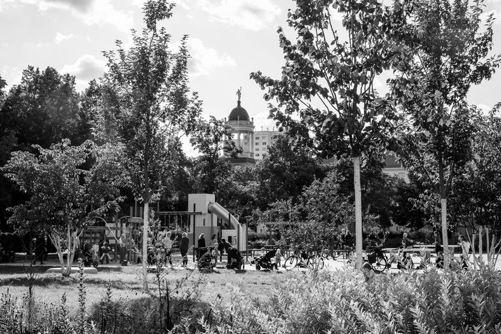 Schwarz-Weiß-Foto zeigt mit Blick durch viel Grün einen belebten Spielplatz, in deren Hintergrund die Kuppel eines Gebäudes empor ragt.
