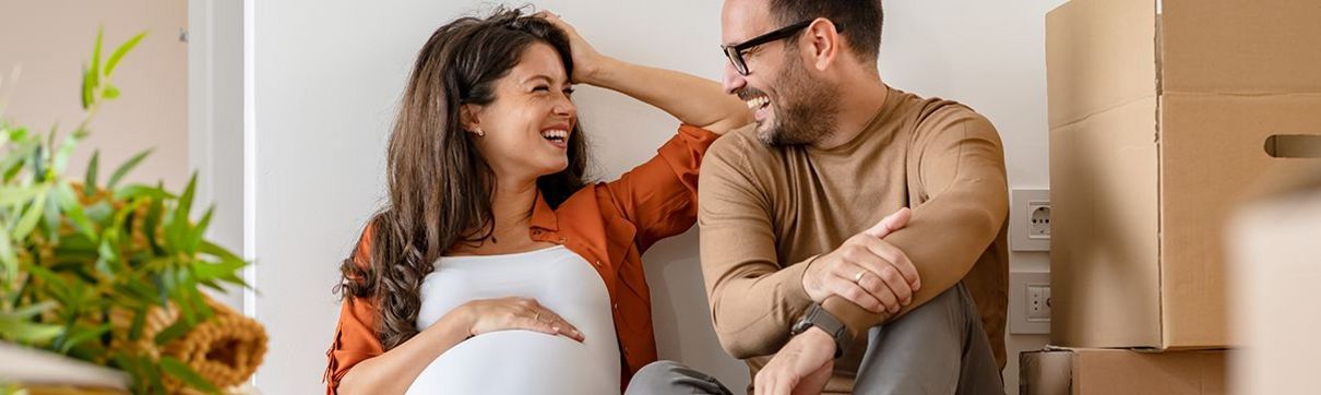 schwangere Frau und Mann sitzen auf dem Boden an die Wand gelehnt und lächeln sich an